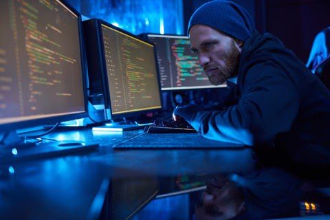 Man wearing beanie is coding with three monitors.