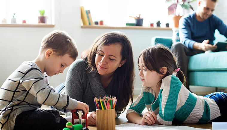 Mom laying on her stomach between her two children watching them draw with husband in the background on sofa to promote importance of life insurance to protect those you love