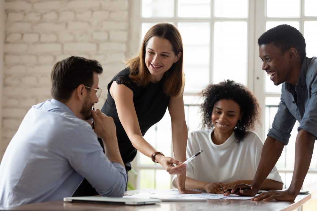 Caucasian woman leader hands pen to man in glasses to sign a contract. Black woman is sitting and observing with a smile on her face Black man is standing, leaning over the table and smiling