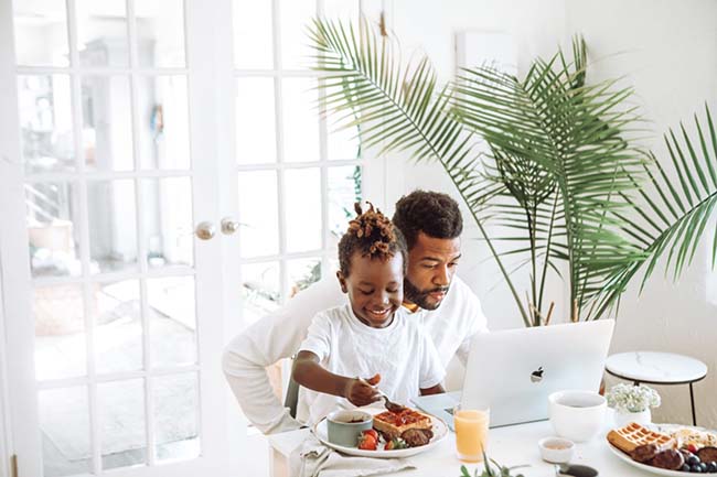 Black man researching life insurance on laptop while eating breakfast and holding his young son